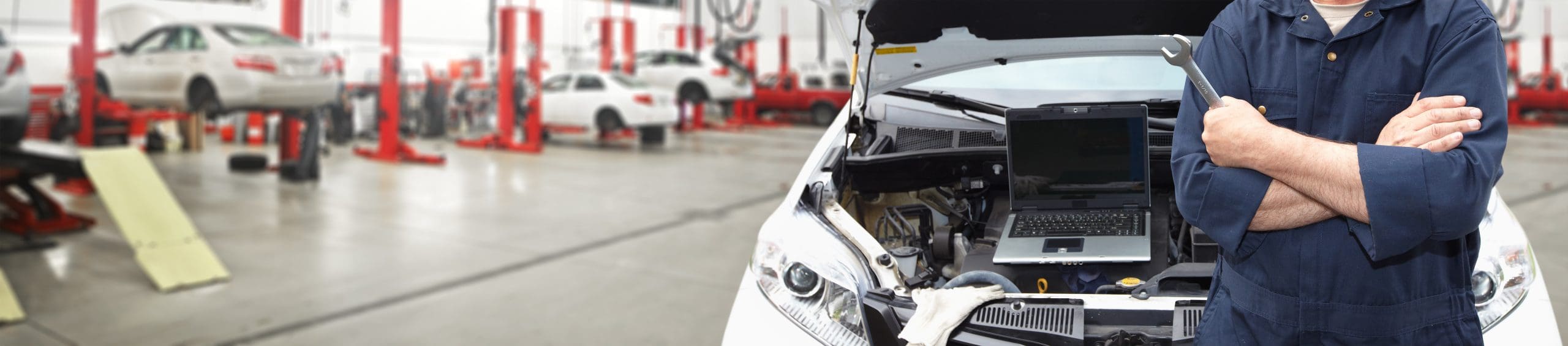 A mechanic in a blue jumpsuit stands with arms crossed in front of a car with its hood open, revealing a laptop placed on the engine. The background shows a busy auto repair shop with several cars raised on hydraulic lifts.