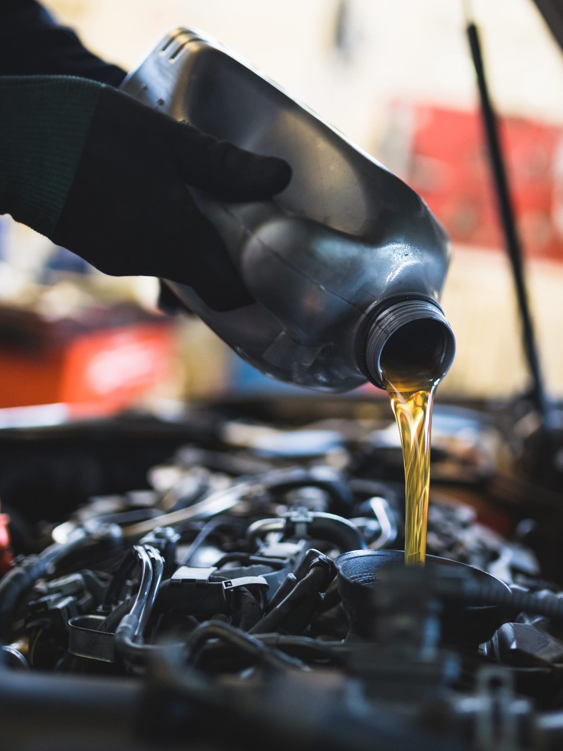 A close-up photo of a person pouring motor oil from a jug into a car's engine. The gloved hand holds the jug steadily over the engine bay, which has various mechanical components visible. The background is slightly blurred, with hints of a workshop setting.