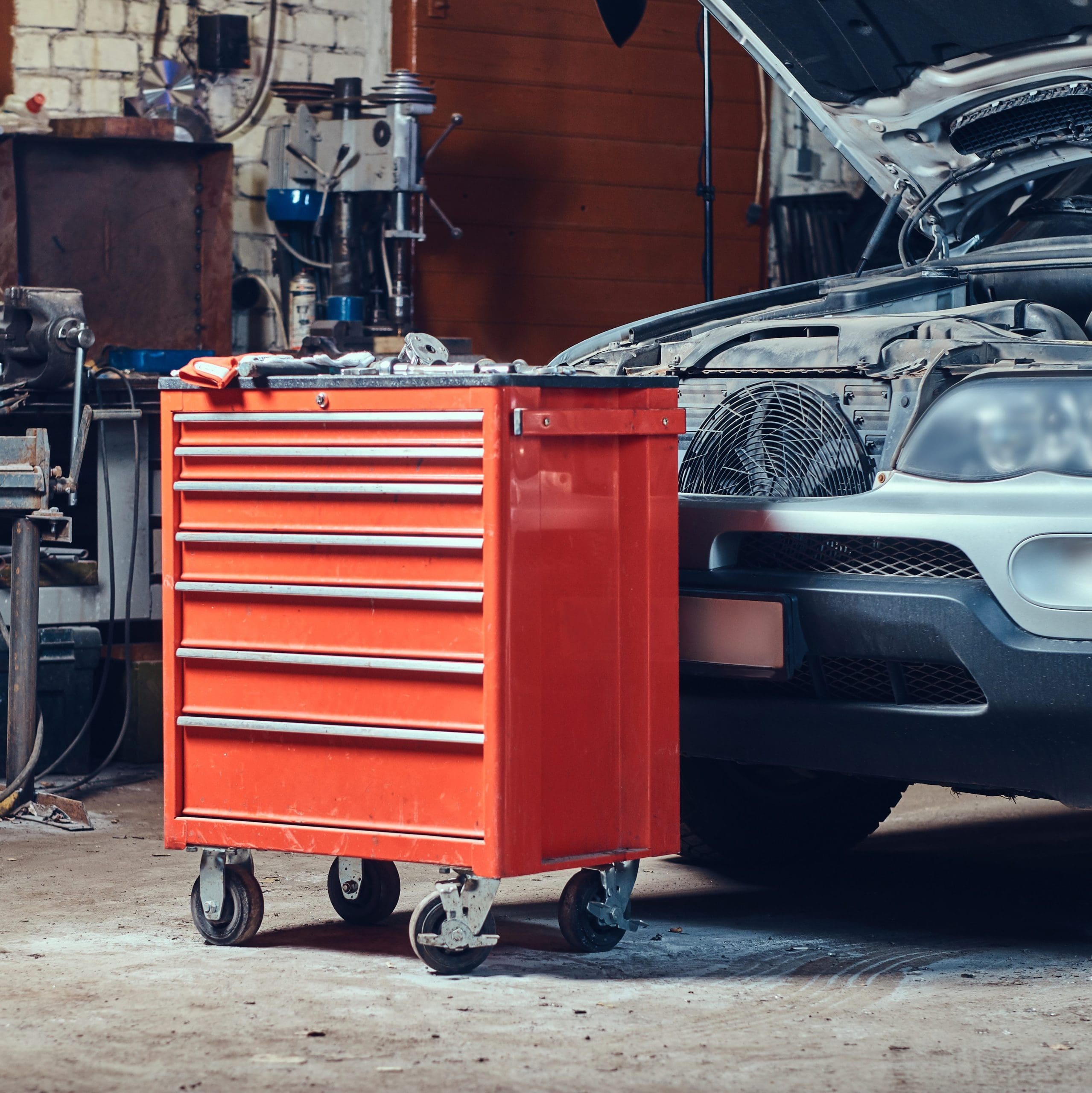 A red tool chest with multiple drawers on wheels stands next to an open car hood in a dimly lit garage or workshop. Various tools are arranged on top of the tool chest. The car and the chest are surrounded by other equipment and tools.