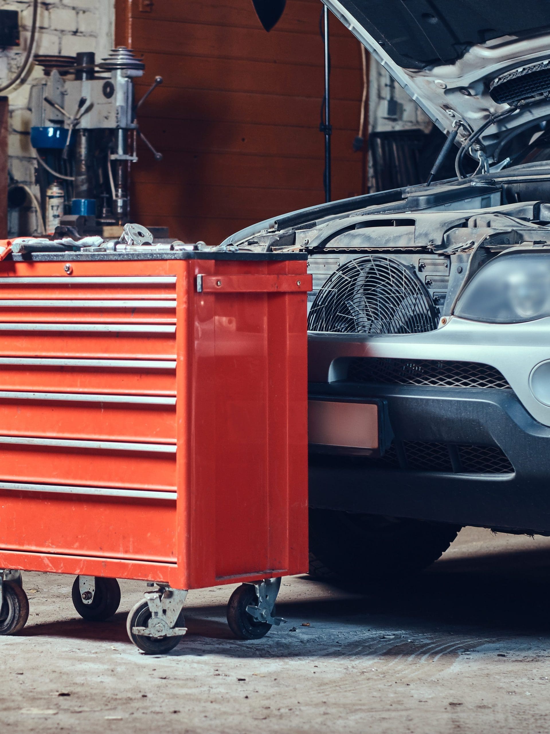 A red tool chest on wheels stands in front of a car with its hood open, parked inside a garage. Various tools and equipment are visible in the background, indicating a workshop or repair setting.