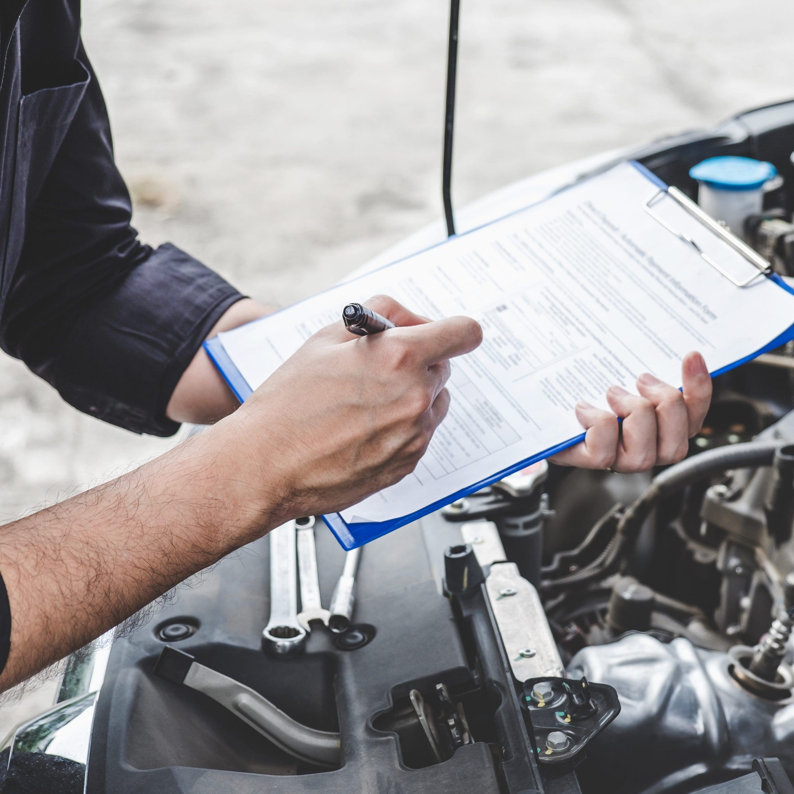 A person holding a blue clipboard and writing on a form with a pen. The person is standing beside an open car hood, suggesting an automotive inspection or repair taking place. Various engine components and tools are visible inside the car hood.