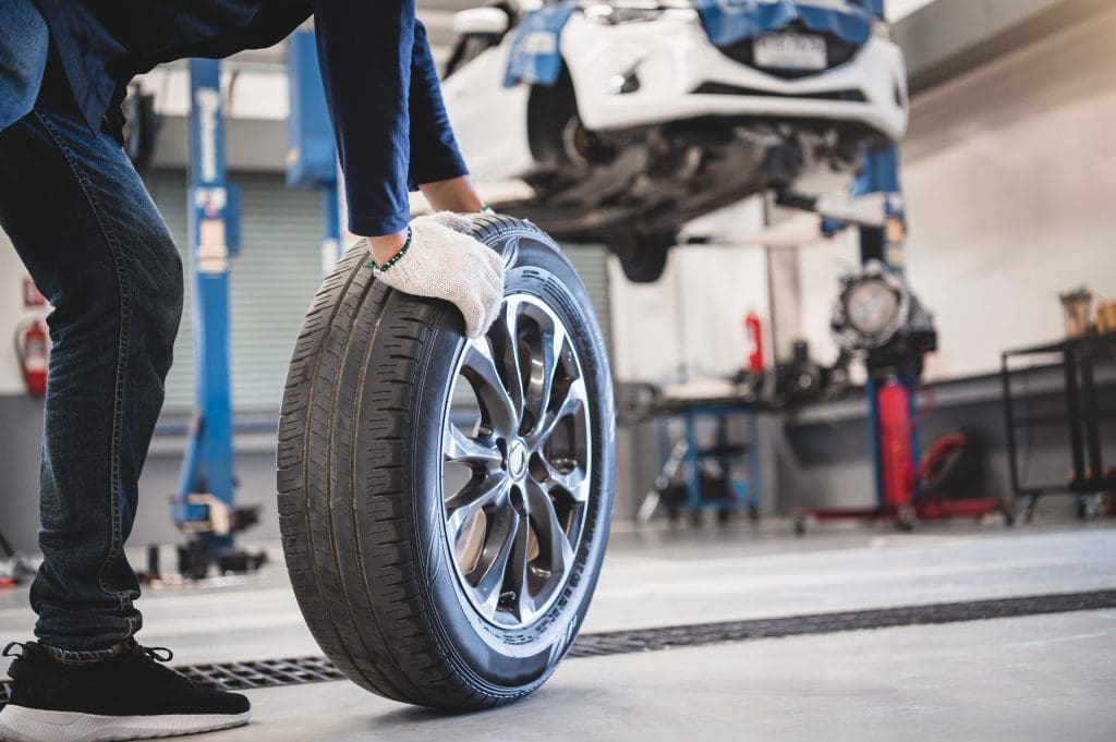 A mechanic with white gloves rolls a car tire in an automotive repair shop. A vehicle is elevated on a lift in the background, indicating maintenance or repair work. The workshop is equipped with various tools and lifts.