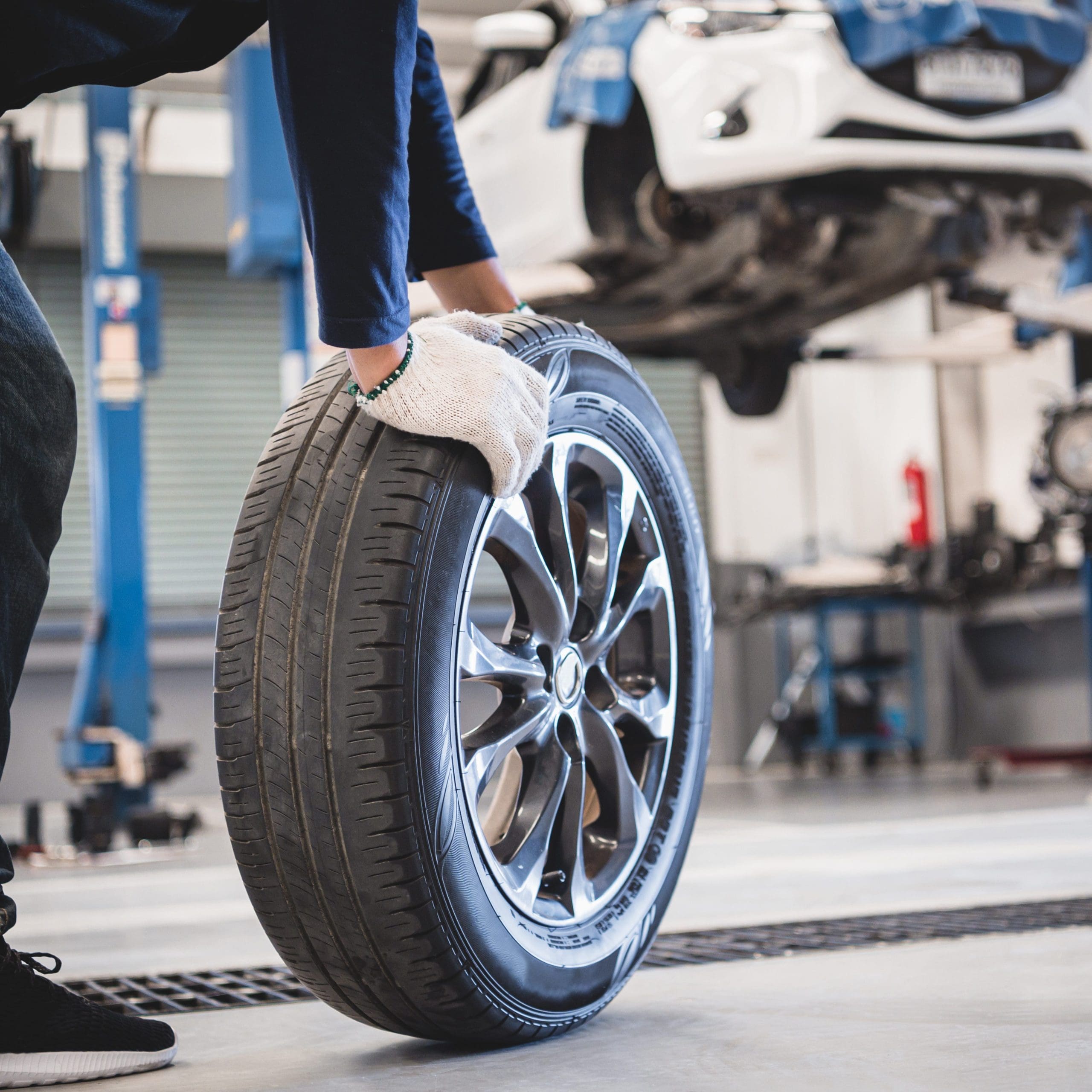 A person wearing gloves rolls a tire on the floor of an automotive repair shop. In the background, a car is lifted on a hydraulic lift, and various tools and equipment are visible.