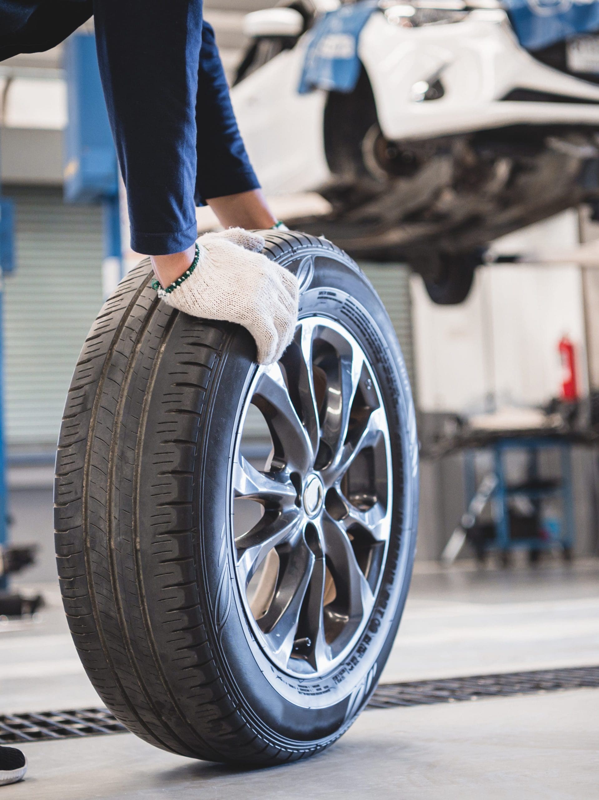 A mechanic wearing gloves is lifting a car tire in a garage. In the background, a white car is elevated on a hydraulic lift, indicating a tire change or maintenance work in progress. The garage is well-lit, with tools and equipment visible in the background.