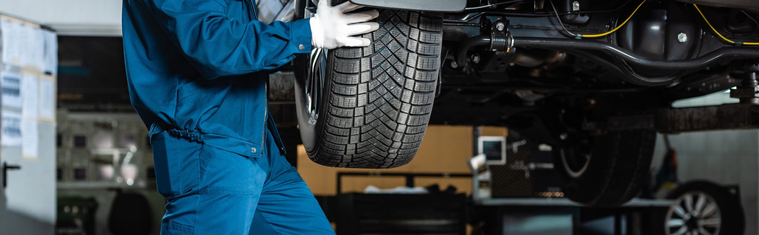 A mechanic in a blue uniform and white gloves is installing a tire onto a vehicle lifted on a car lift in a garage. The scene shows a detailed close-up of the tire and the lower half of the mechanic's body.