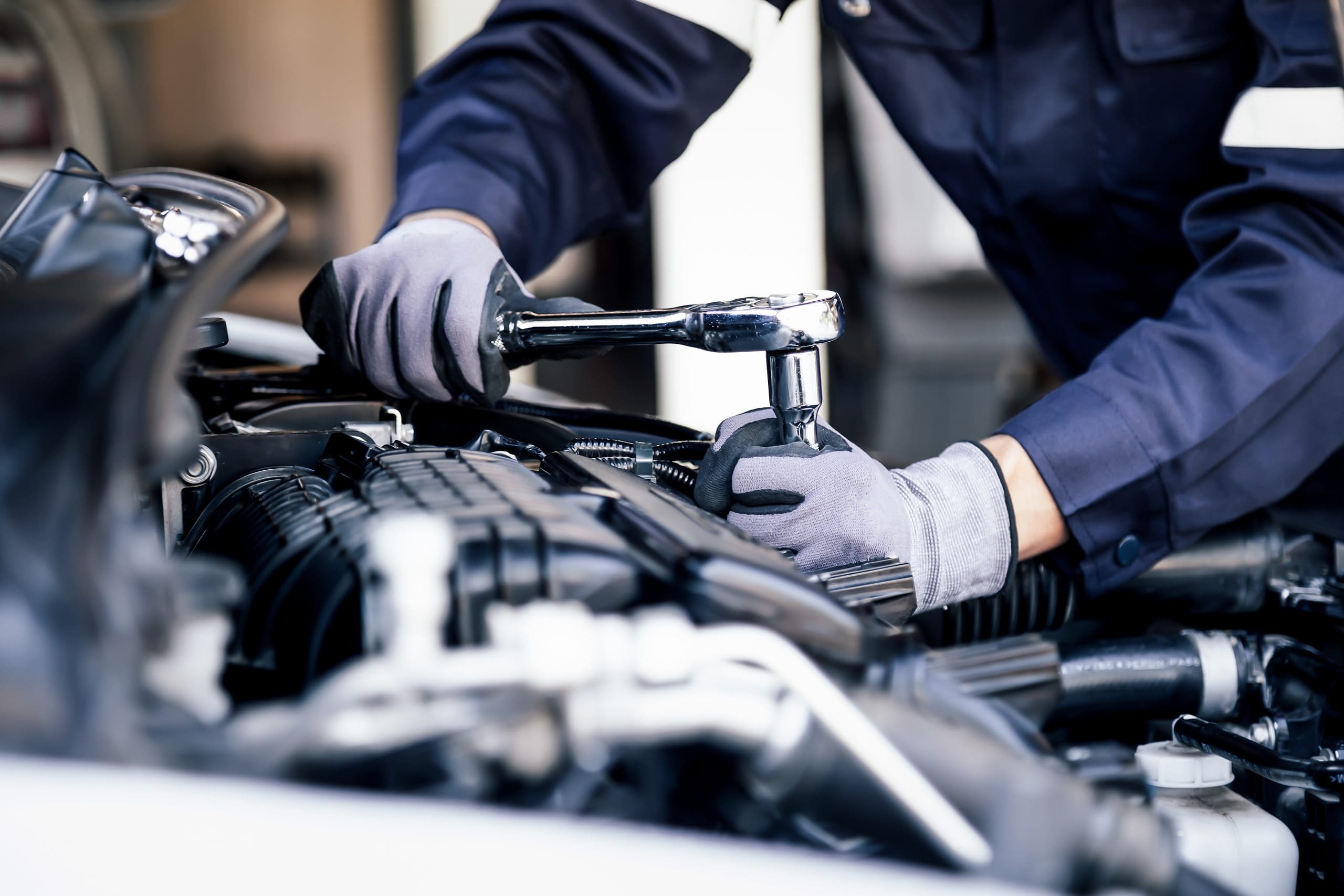 A mechanic wearing gloves and a blue jumpsuit uses a ratchet wrench to work on a car's engine. The close-up shot emphasizes the mechanic's hands and the tools, highlighting the detailed work involved in car maintenance.