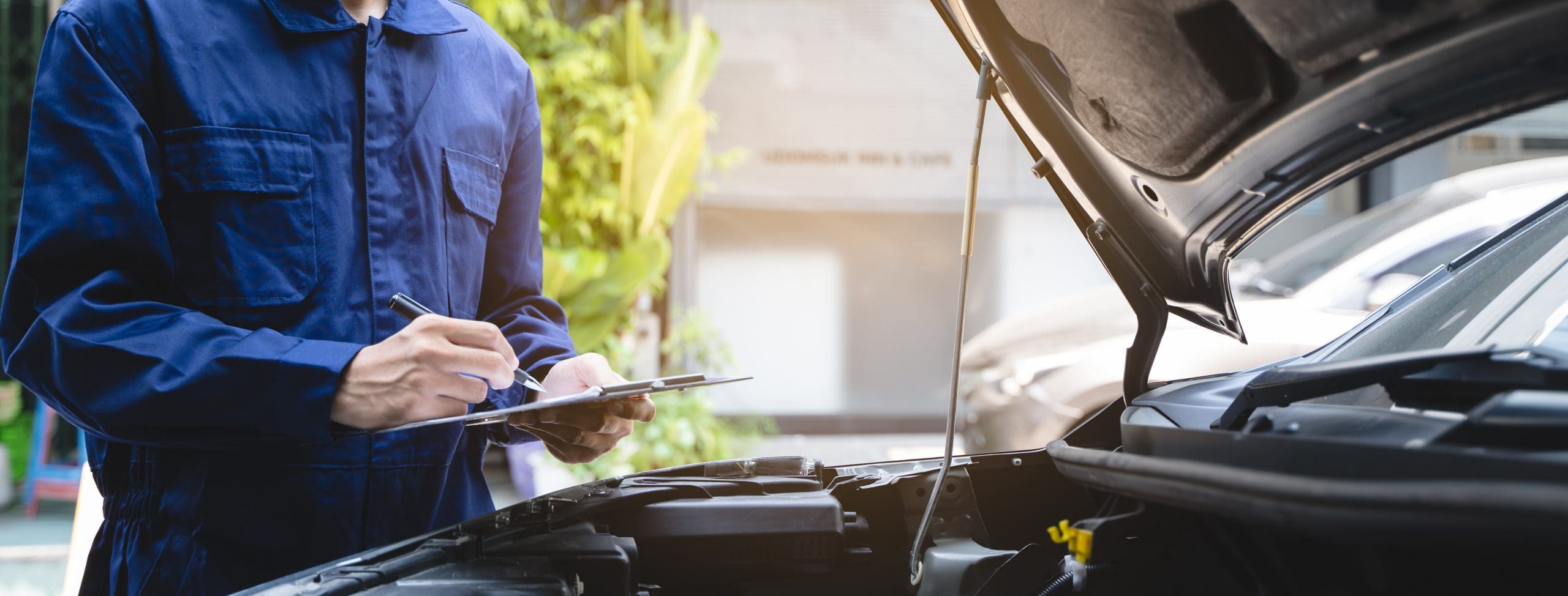 A mechanic in a blue uniform holds a clipboard and writes on it while inspecting a car with its hood open. The background shows greenery and a blurred building.