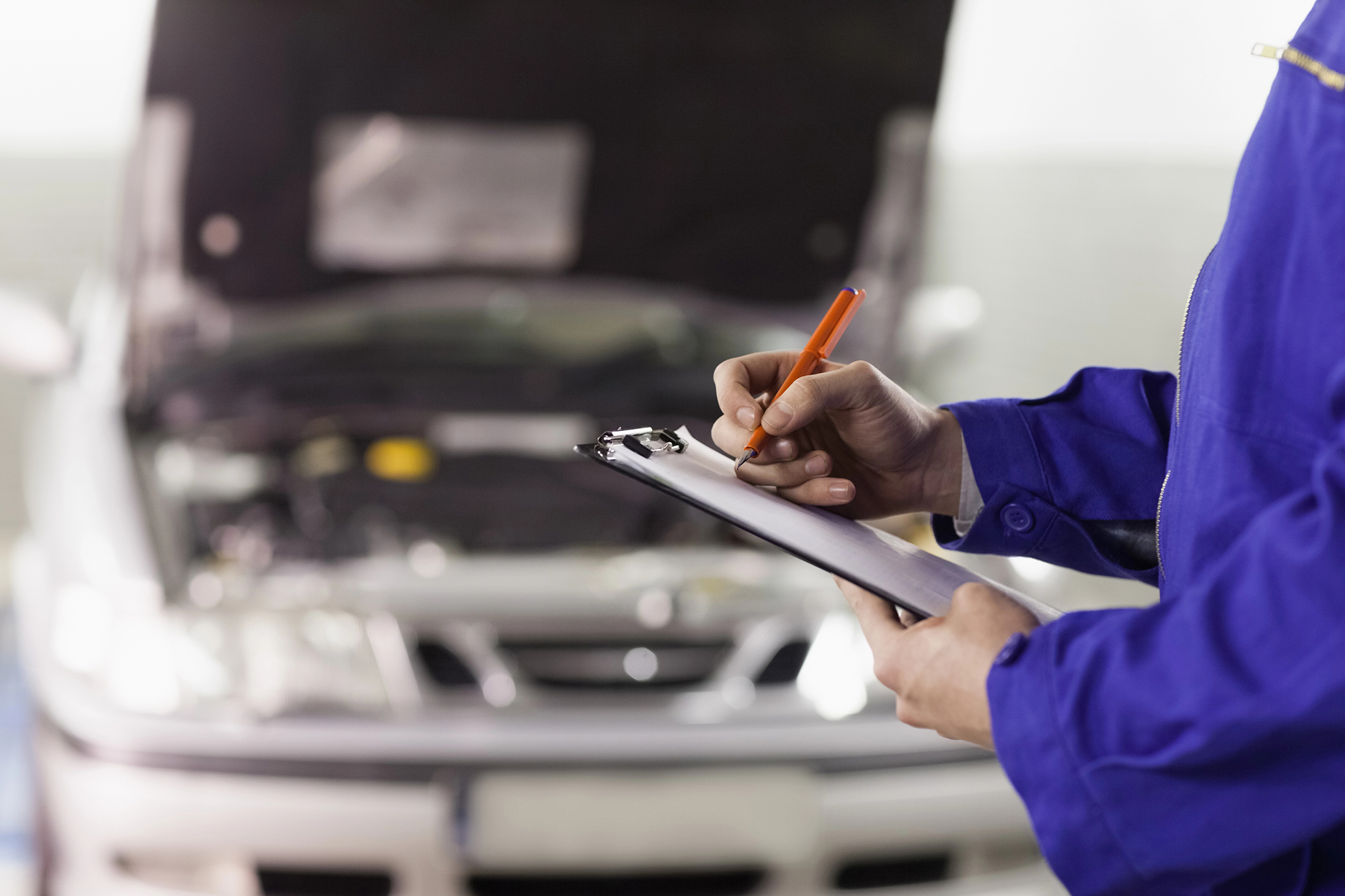 A mechanic in a blue uniform is writing on a clipboard with an orange pen. Behind them, the hood of a silver car is open, indicating that the car is being inspected or repaired. The background is slightly blurred, focusing attention on the mechanic's actions.