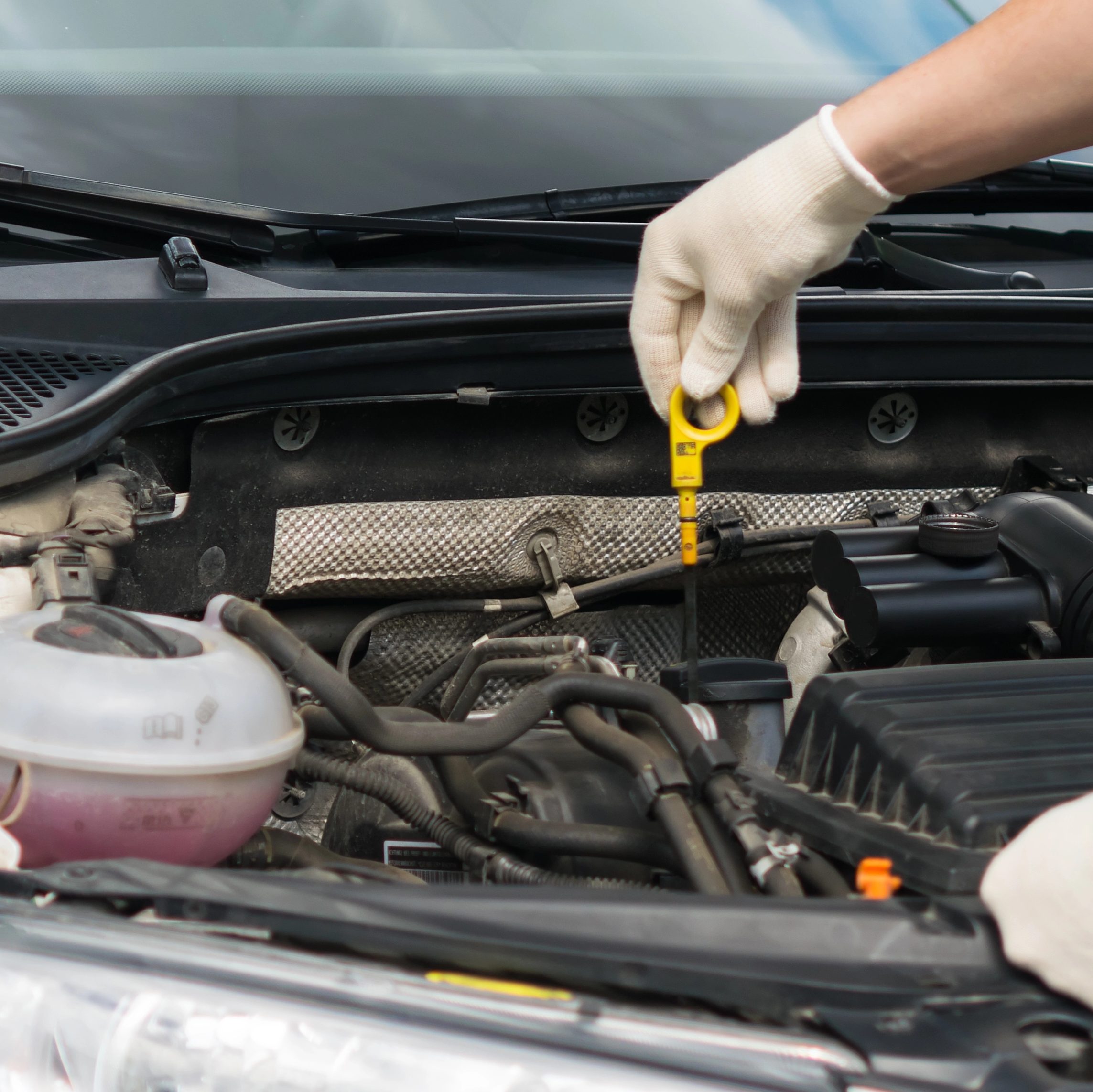 A person wearing white gloves checks the oil level of a car engine using a dipstick. The hood of the car is open, revealing various engine components and the engine bay's interior. The dipstick is marked for measuring oil levels.