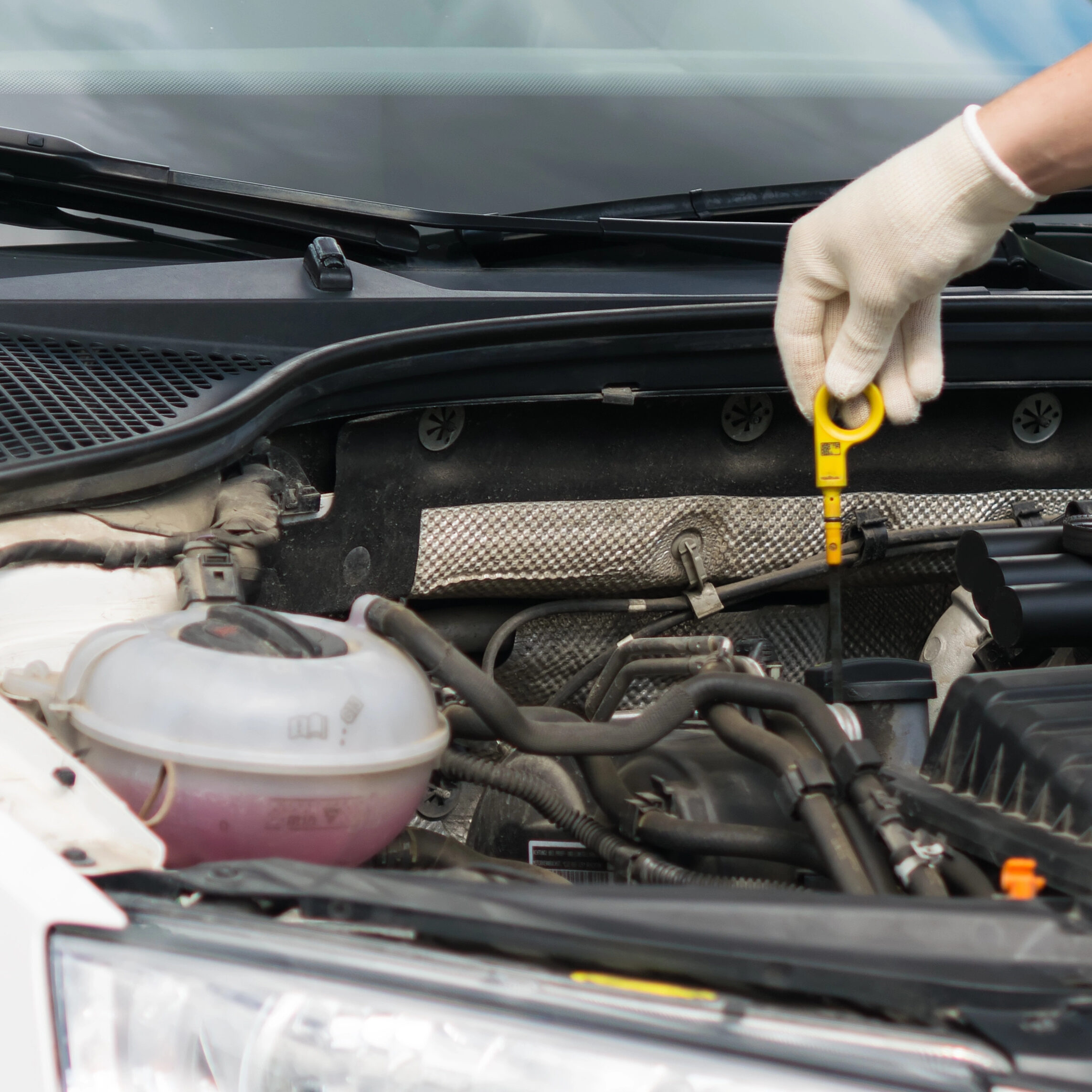 A person wearing white gloves checks the oil level in a car engine using a yellow dipstick. The car's hood is open, revealing various engine components, with the dipstick being pulled out for inspection.