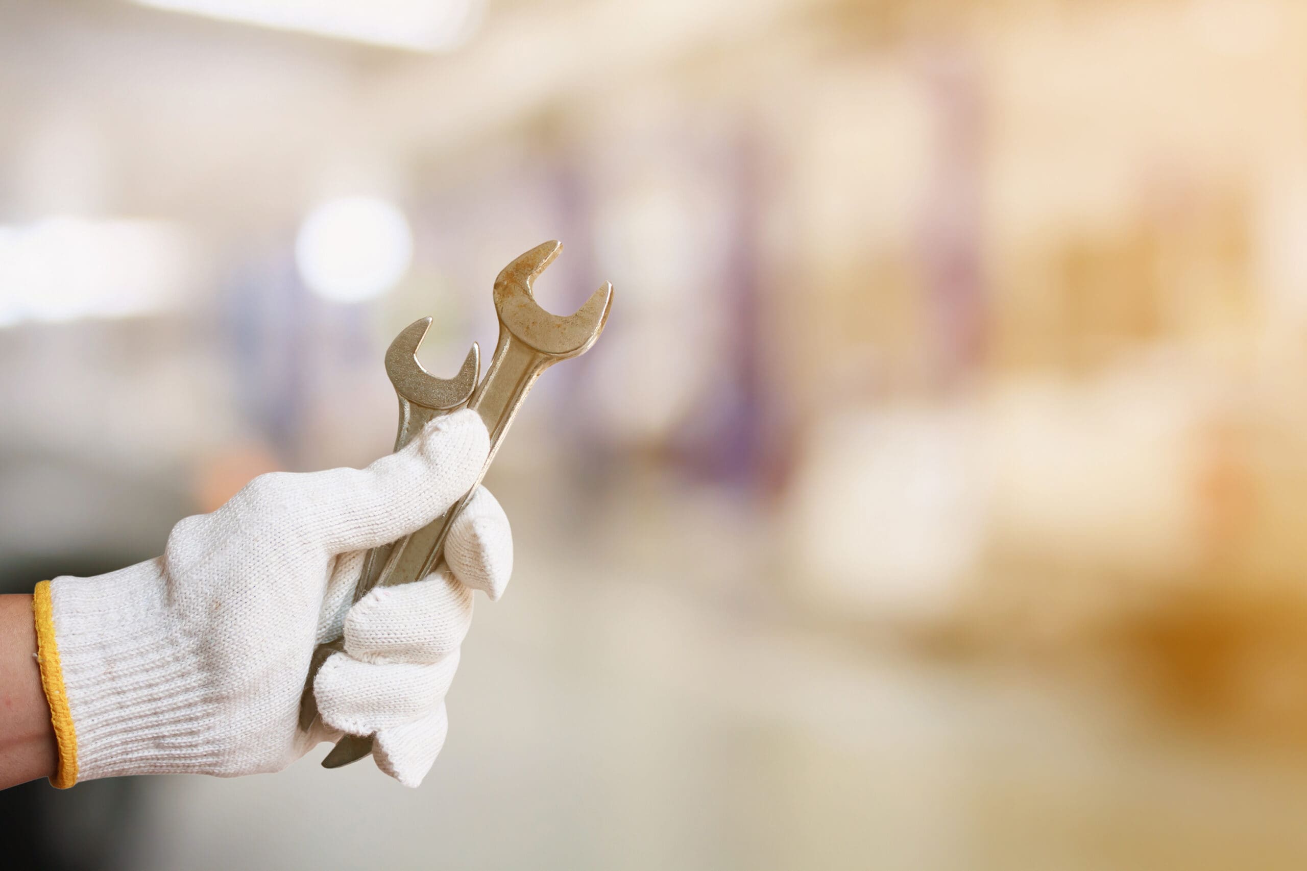 A hand wearing a white work glove holds two wrenches against a blurred background, which appears to be a workshop or industrial setting. The image conveys a sense of manual work and repair.