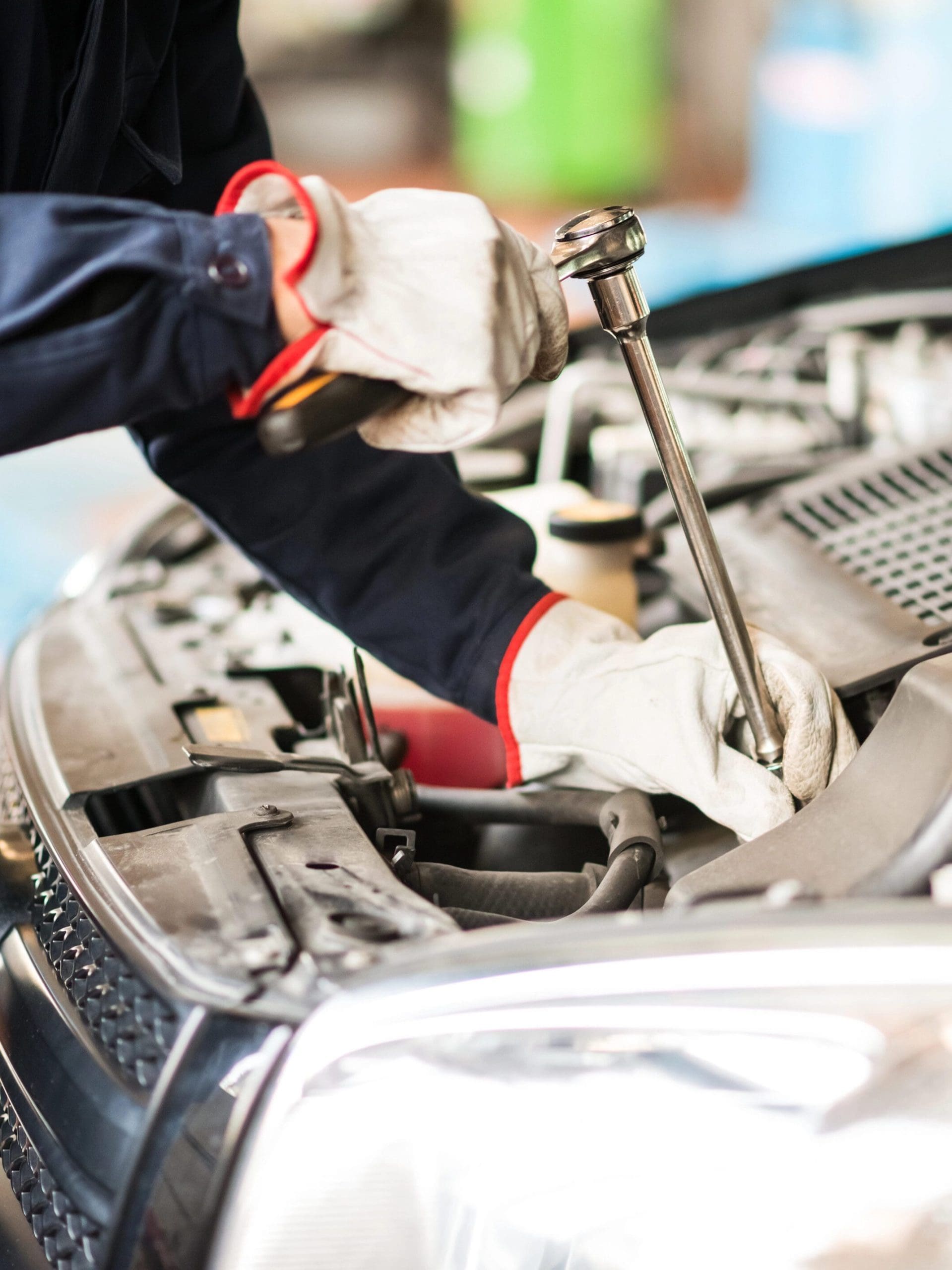 A mechanic wearing protective gloves is using a ratchet wrench to work on a car engine. The car's hood is open, revealing various components of the engine. The mechanic's focus is on a specific part, indicating detailed and careful work.