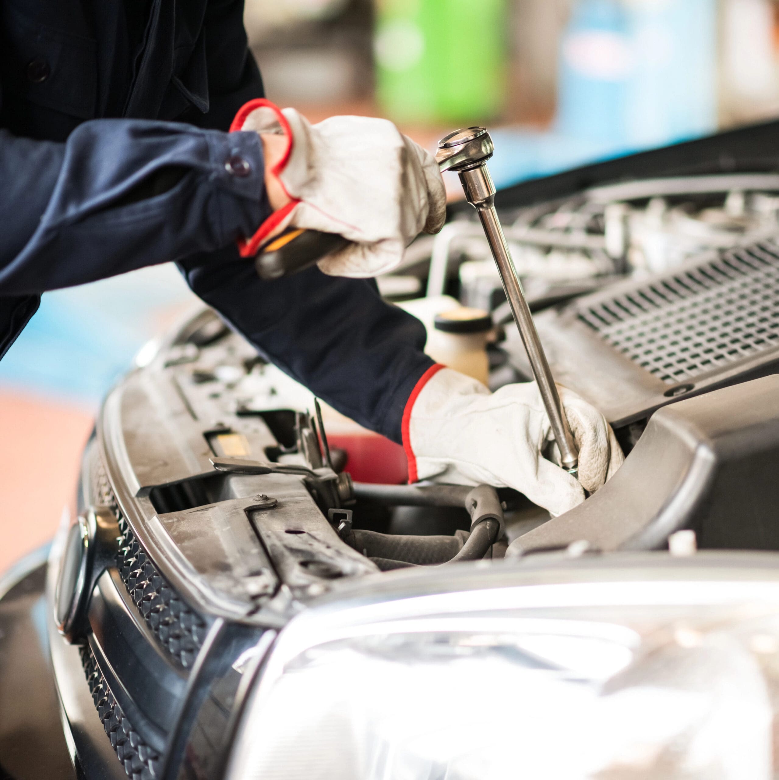 A mechanic wearing gloves and a navy uniform with red cuffs uses a wrench to work on the engine of a car with the hood open. The image focuses on the hands and tool, part of the vehicle's front grille and engine area visible.