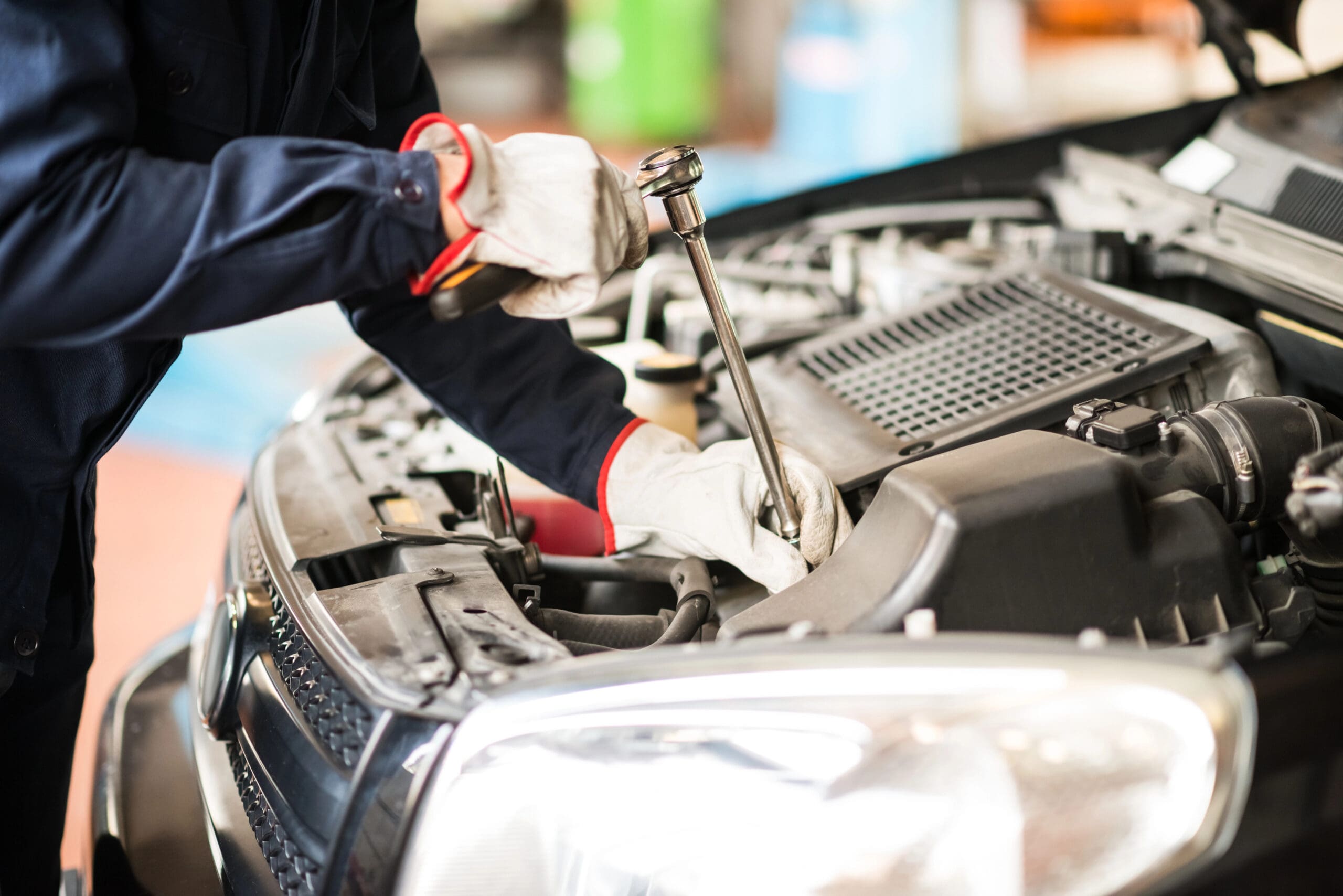 A mechanic wearing gloves and a dark uniform is tightening a bolt under the hood of a car with a wrench. The engine components and front grille of the car are visible, indicating a vehicle maintenance or repair scenario.