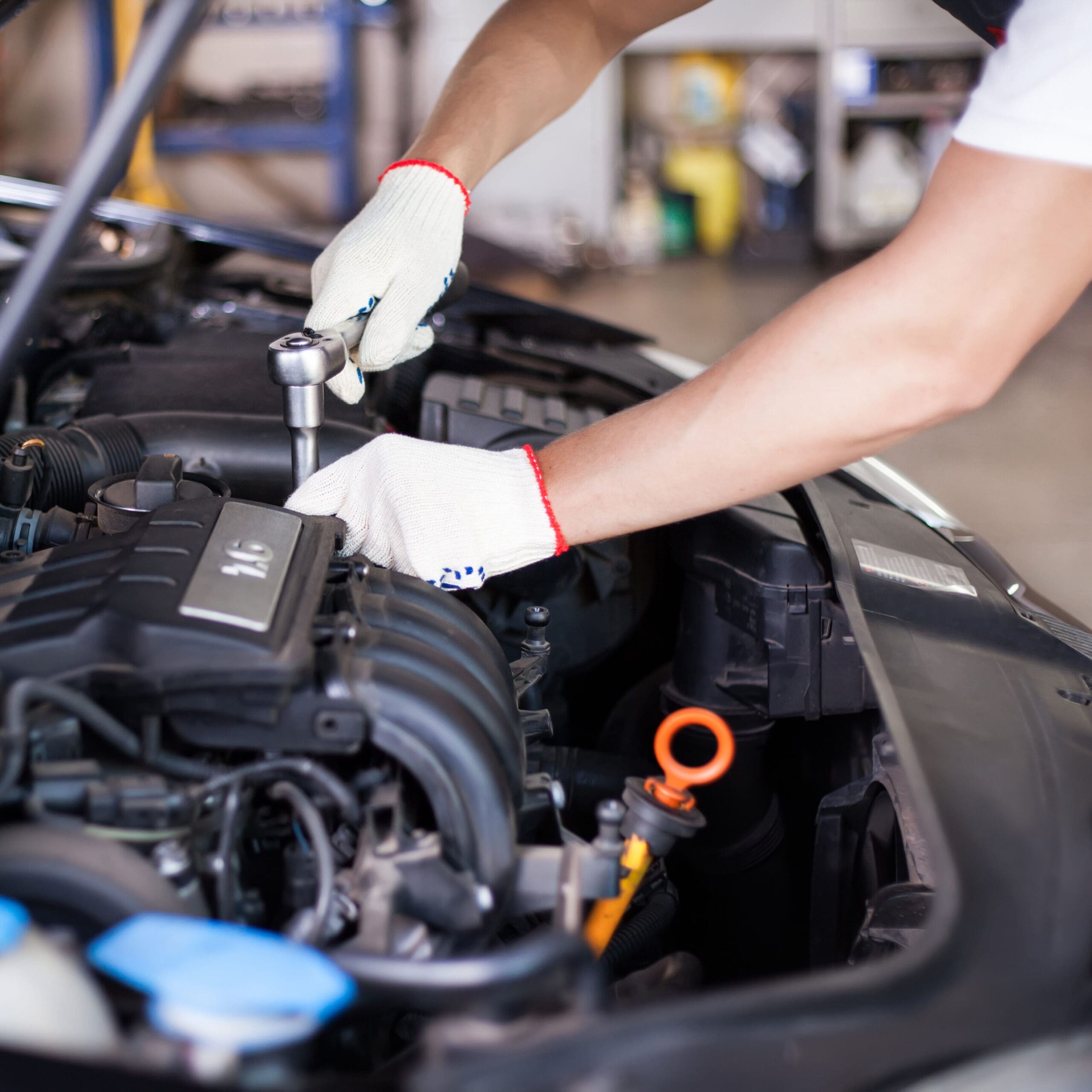 A mechanic in white gloves uses a ratchet wrench to work on the engine of a car in a workshop. The car's hood is open, providing a view of various engine components. The background shows a blurred workshop environment with tools and equipment.