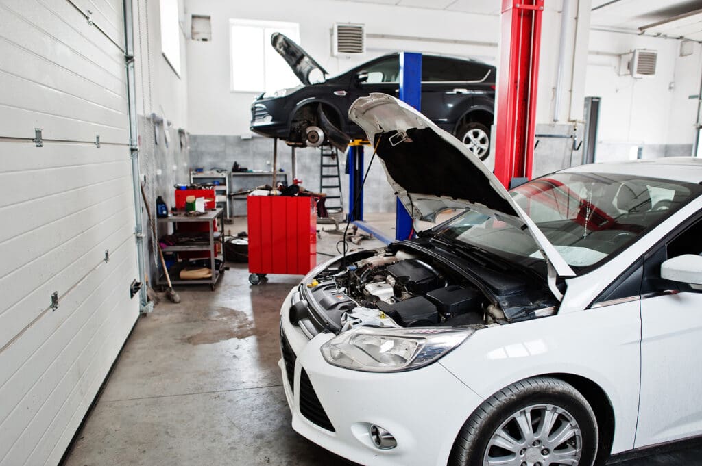 Two cars are being serviced in an auto repair shop. The nearest car, a white sedan, has its hood open, and a black car is lifted on a hydraulic lift in the background. Various tools and equipment are scattered around the garage.