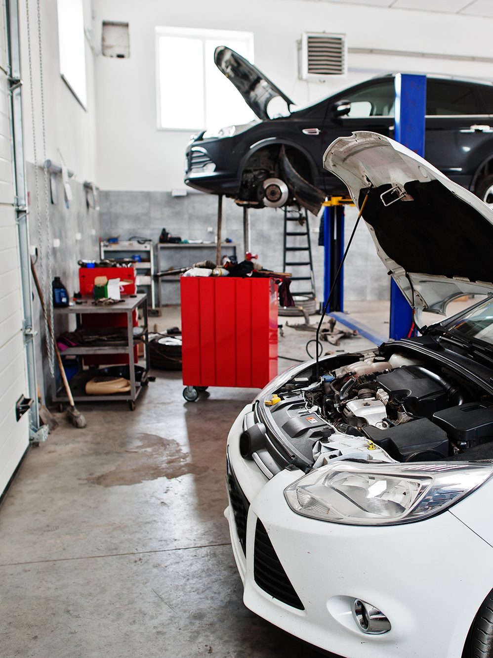 A car repair shop with two vehicles being worked on. One car is raised on a hydraulic lift with its hood open and tires removed, while the other car is on the ground with its hood open. Various tools and equipment are scattered around the clean, well-lit garage.