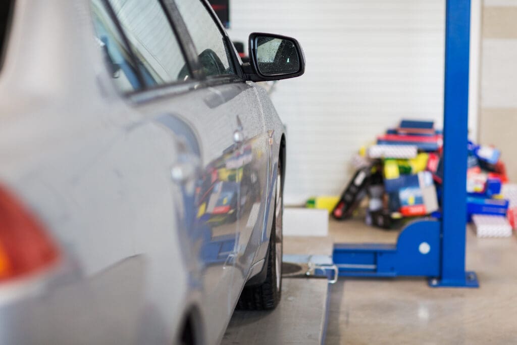 A silver car is positioned at an angle on a hydraulic lift in an automotive repair shop. The background shows various colorful boxes and automotive parts stacked and scattered near a white wall. The car's side mirror is visible and reflects part of the shop.