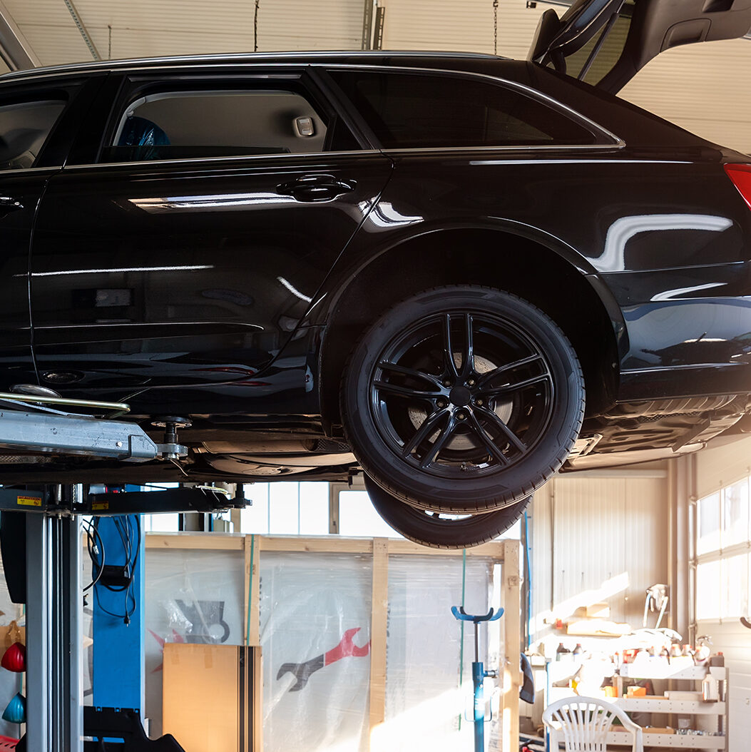 A black car is elevated on a lift inside an auto repair shop. The rear door is open, and the shop is lit by natural sunlight streaming through the windows. Various tools and equipment are visible in the background.