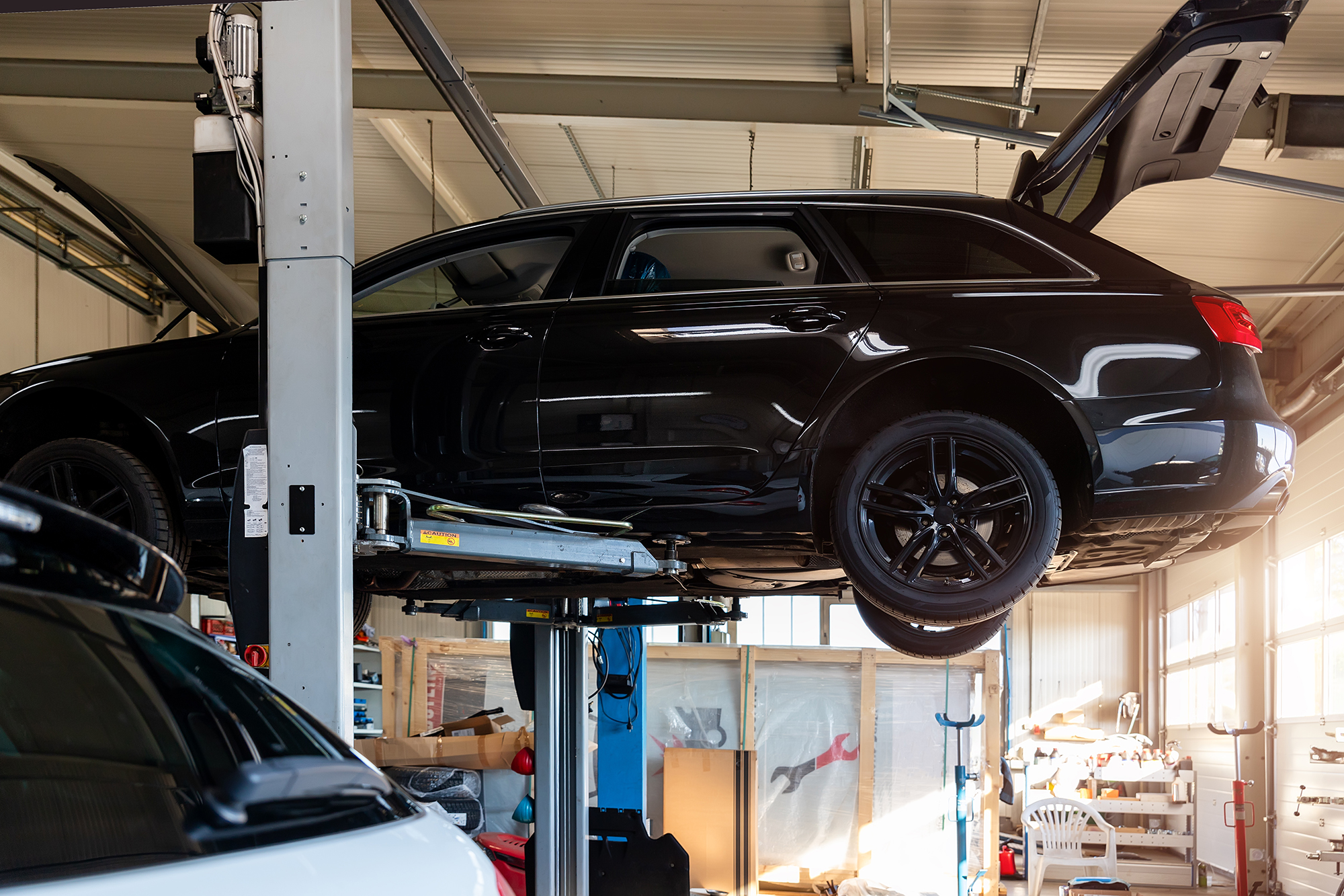 A black car is elevated on a hydraulic lift in a garage, with its trunk open. The surrounding area is filled with various tools and equipment, indicating an auto repair or maintenance setting. Sunlight streams in through the windows, illuminating the workspace.