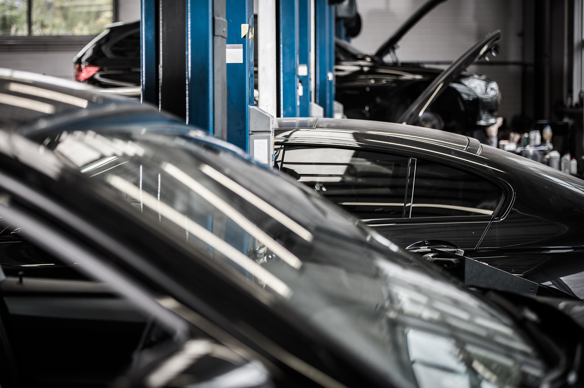 A vehicle repair shop with several cars being worked on. Cars are parked in a line, some with their hoods open. Blue lifting equipment supports the vehicles. The image focuses on the side view of dark-colored cars and the workshop environment.