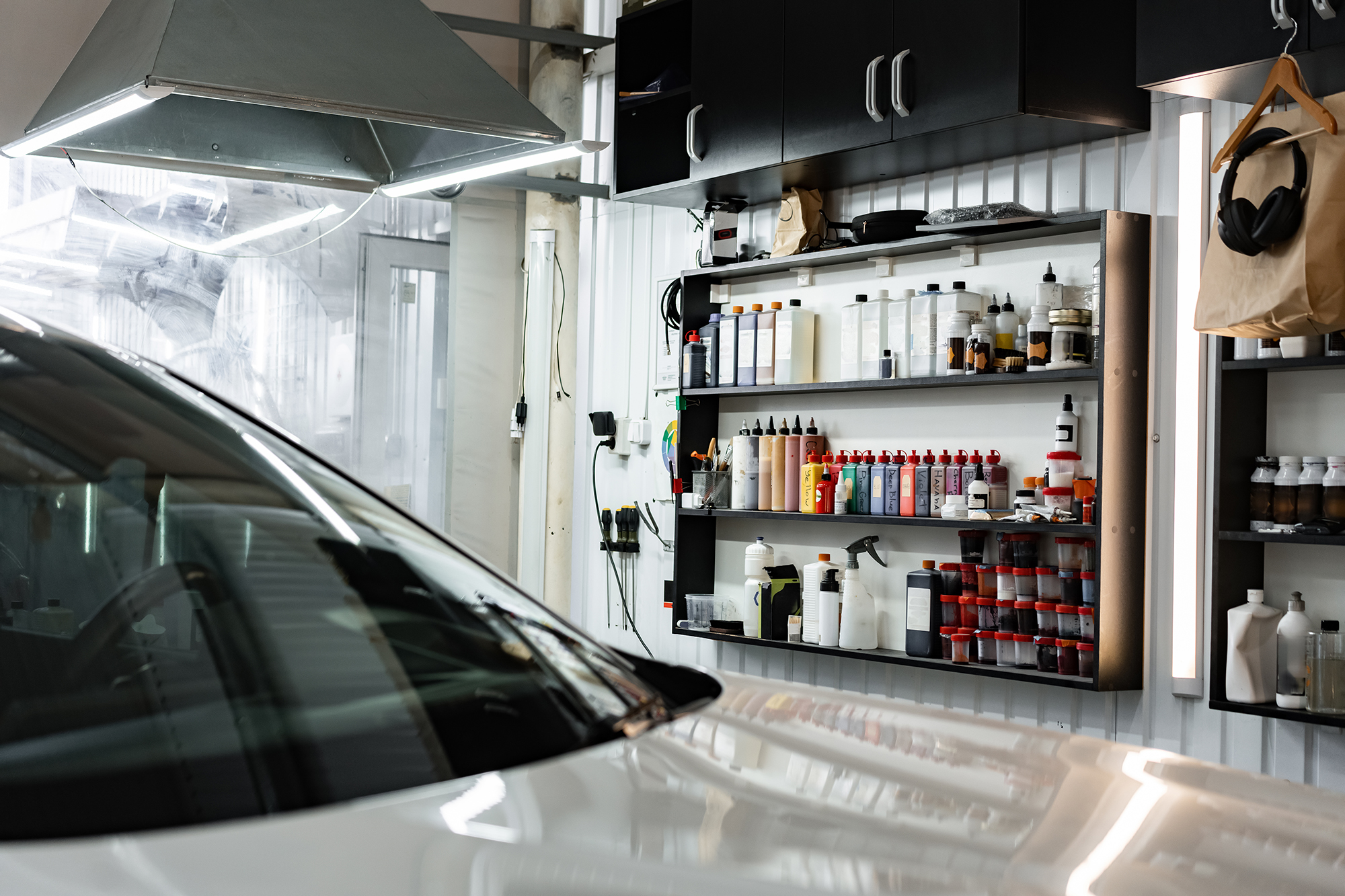 A well-organized car detailing workshop with a variety of cleaning and polishing products neatly arranged on shelves. In the foreground, the glossy front end of a white car is partially visible. The workspace is brightly lit and clean.