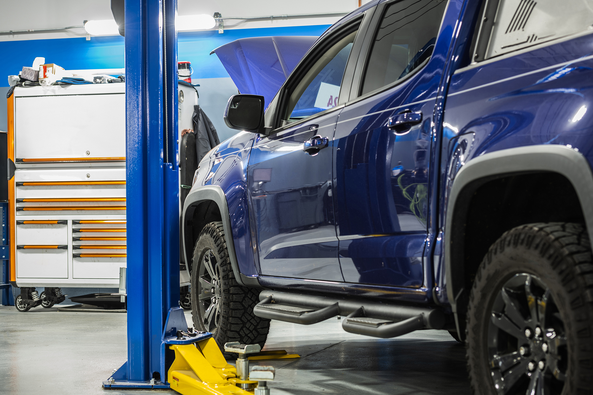 A blue pickup truck is lifted on a hydraulic car lift inside an auto repair shop. A toolbox with drawers is on the left side, and the hood of another vehicle is open in the background. The shop has a clean, organized appearance.