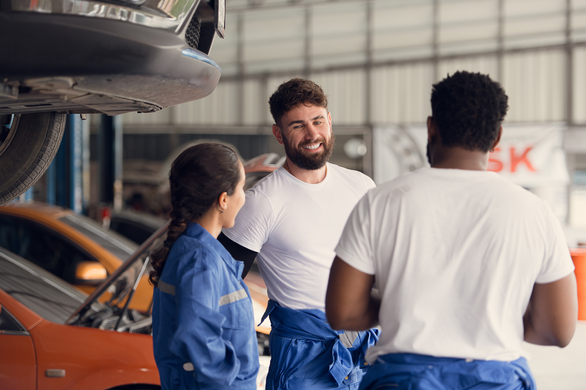 Three mechanics, two men and one woman, stand in a workshop next to raised cars. They are wearing blue overalls and white shirts. The mechanic with the beard is talking and smiling. The surrounding space showcases a garage environment with tools and equipment.