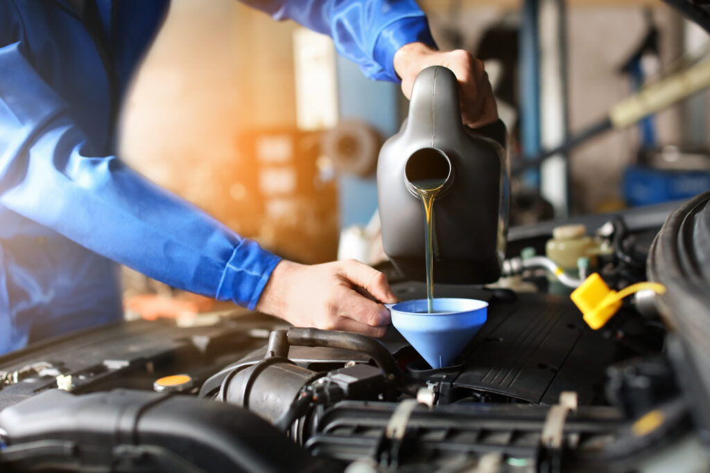 A person in a blue uniform pours engine oil from a black container into a car engine using a blue funnel, with the car hood open in a garage setting.