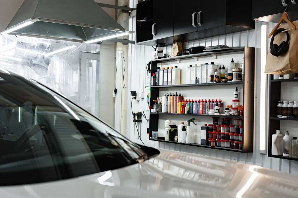 A car is parked inside a garage or workshop with shelves filled with bottles, spray cans, and cleaning supplies against the wall. Overhead cabinets and a large ventilation hood are also visible.